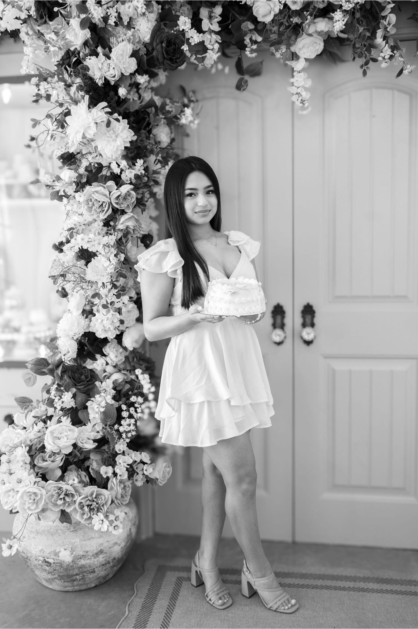 A young woman with long dark hair, wearing a white dress and heels, holding a cake and standing in front of a floral arch ...